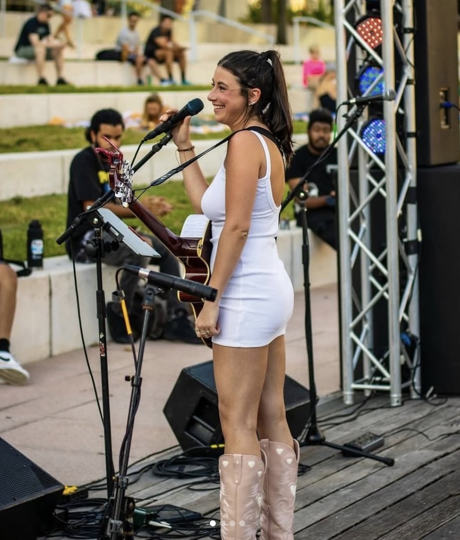 Leanne McGroary performing live with her guitar, singing into a microphone in a white dress and cowboy boots