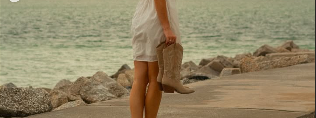 Leanne McGroary in a white dress walking on a rocky jetty by the ocean, carrying cowboy boots