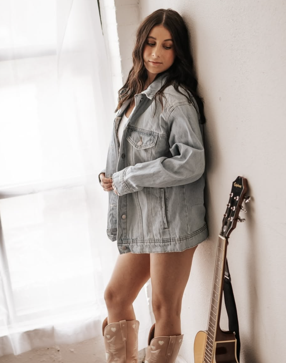 Leanne in a denim jacket leaning against a wall with her guitar